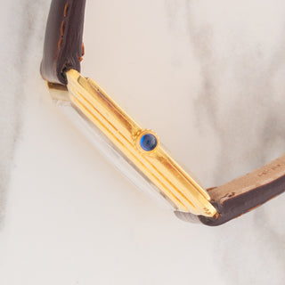 Side profile view showcasing the stepped gold case sides and blue sapphire cabochon winding crown on a Cartier Tank