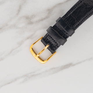 Close-up view of the gold-toned pin buckle attached to a black crocodile leather watch strap