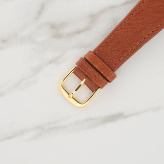 Close-up view of the gold-toned pin buckle attached to a textured brown leather watch strap