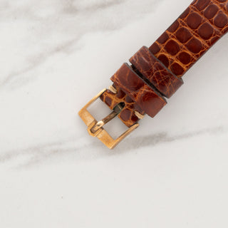 Close-up view of the gold-toned pin buckle attached to a brown crocodile-grain leather watch strap