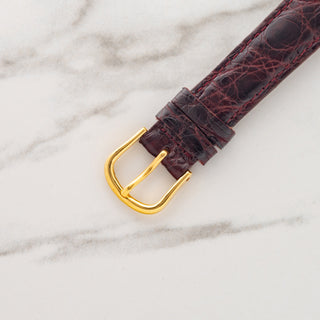 Close-up view of the gold-toned pin buckle attached to a glossy burgundy alligator leather watch strap